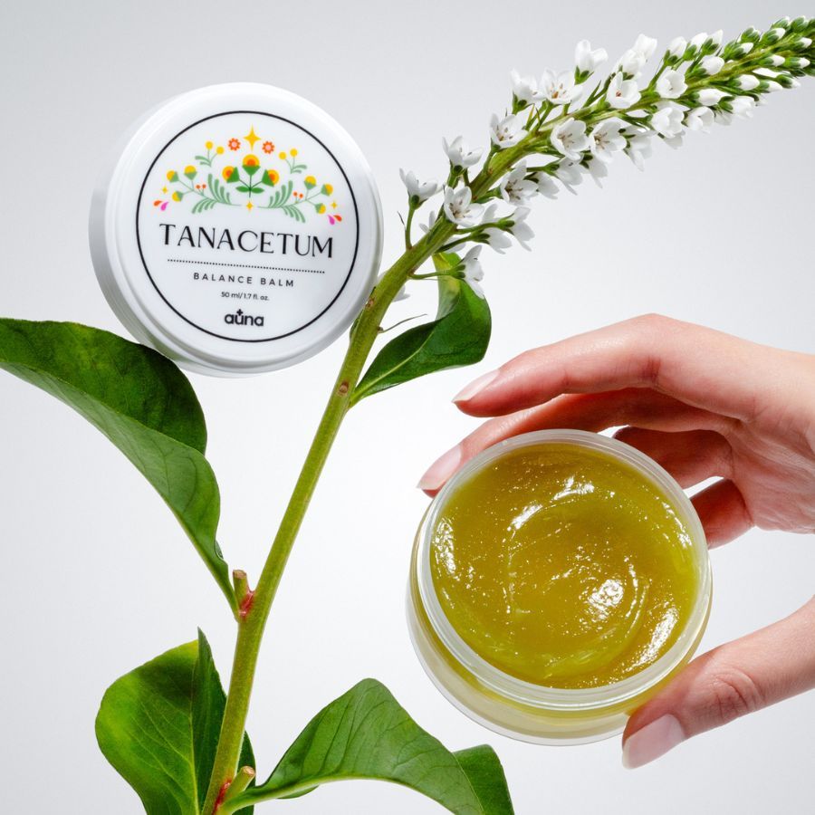 Geöffnete Dose mit grünlichem Balsam, neben einer Hand und einer Pflanze. Weißer Deckel mit floralem Etikett. Text: TANACETUM BALANCE BALM, auna.