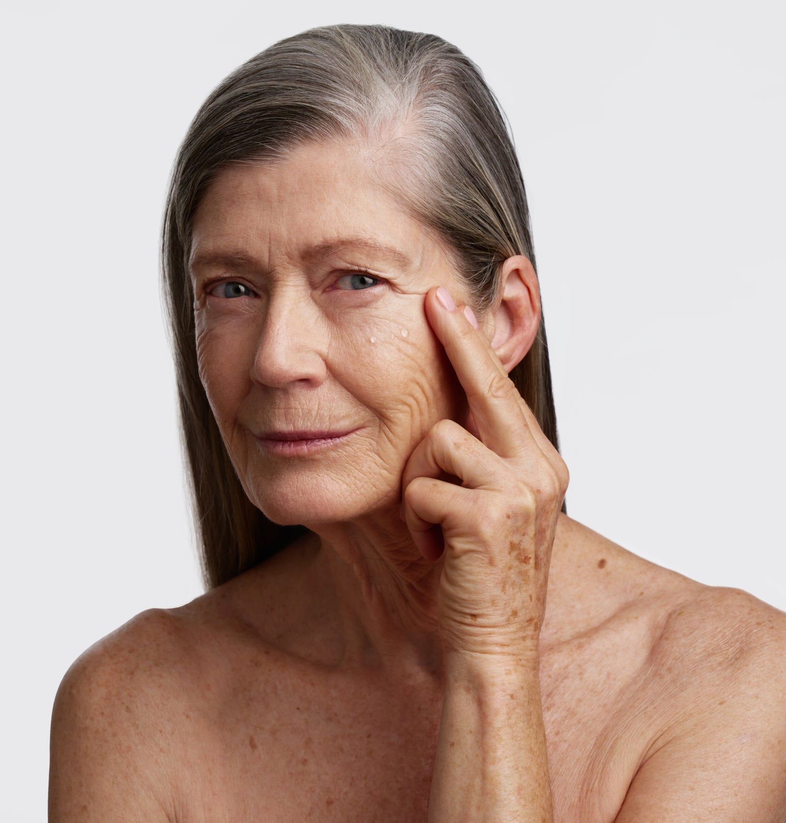 Ältere Frau berührt Gesicht. Haut mit Tröpfchen. Graue Haare, freier Oberkörper. Blick in die Kamera.