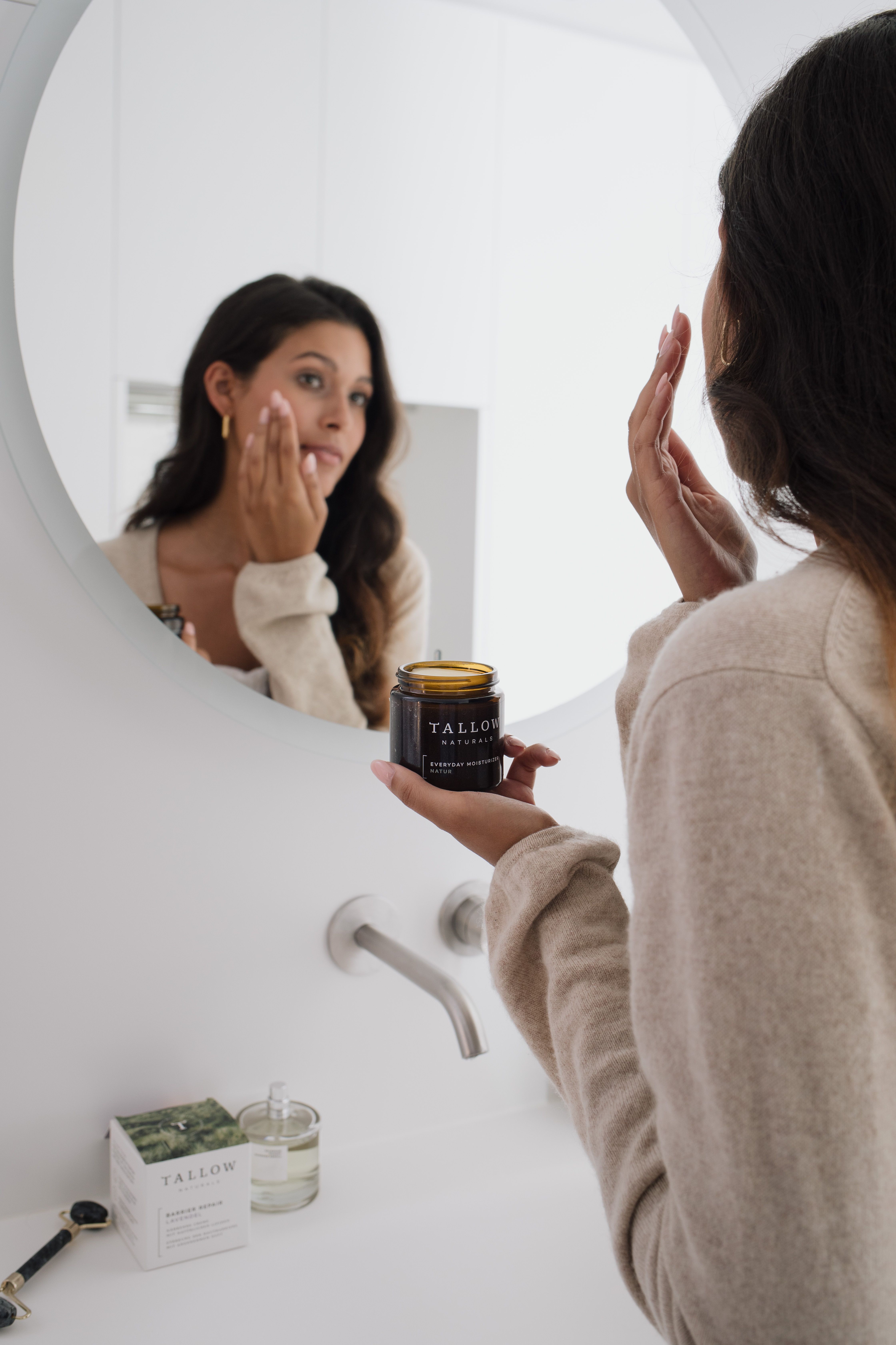 Frau betrachtet sich im Spiegel. Sie hält ein braunes Glas mit schwarzem Deckel. Aufschrift: TALLOW NATURALS, BARRIER REPAIR LAVENDEL.
