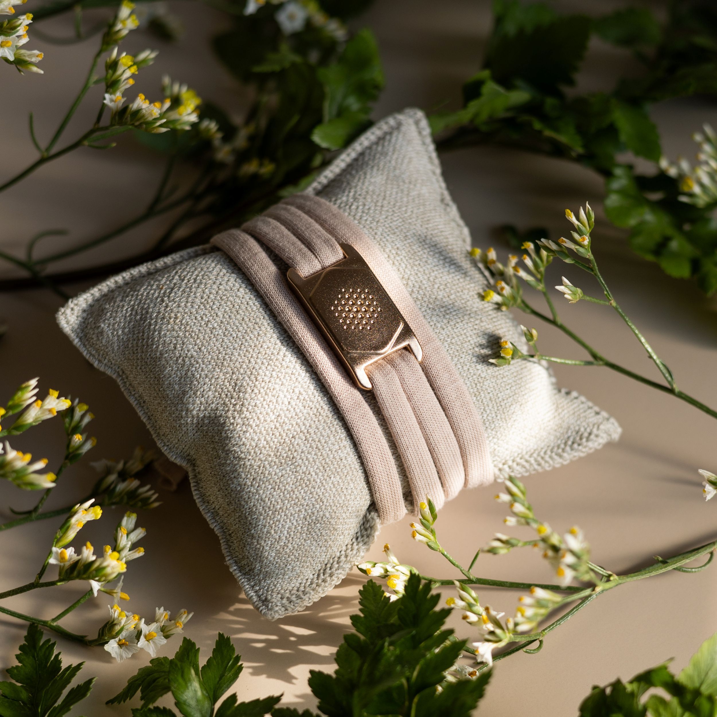 Beiges Armband mit goldfarbenem Verschluss auf einem grauen Kissen, umgeben von grünen Blättern und Blüten.