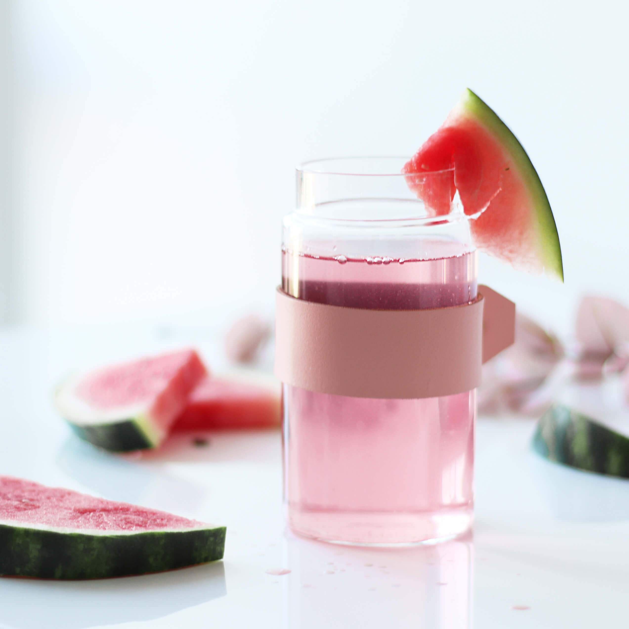 Glas mit rosa Getränk, Wassermelonenstück. Rosa Halterung. Wassermelonenstücke im Hintergrund.