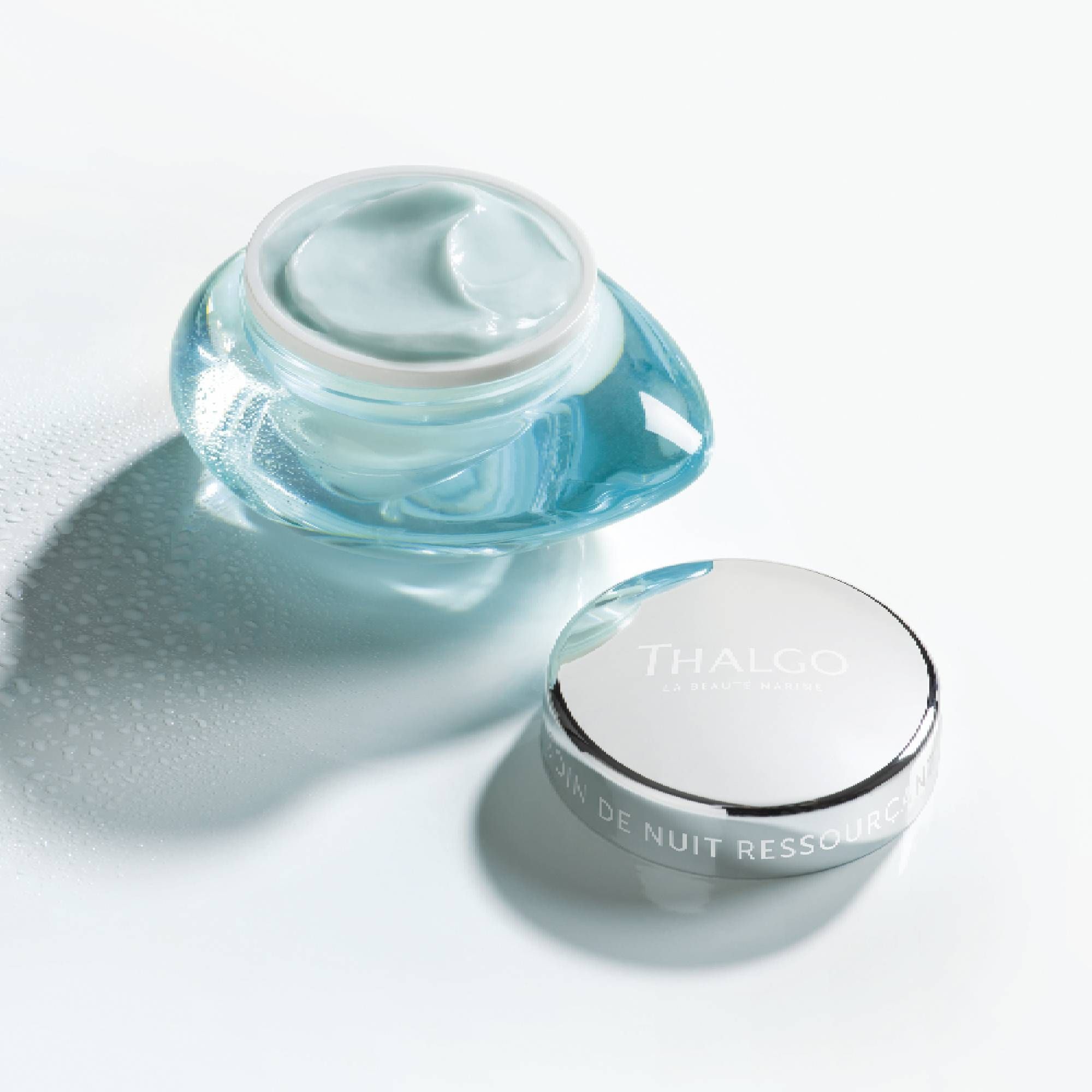 Geöffnete, hellblaue Creme in einem Glasbehälter mit silbernem Deckel. Deckel daneben. Aufschrift: THALGO, SOIN DE NUIT RESSOURCANT.