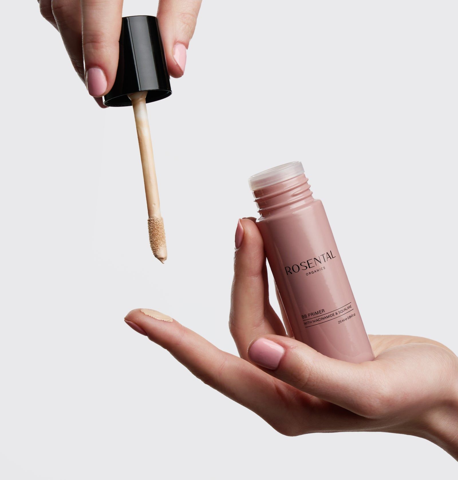 Hände halten eine BB Primer-Flasche und einen Applikator. Flasche rosa, schwarzer Deckel. Applikator hell.