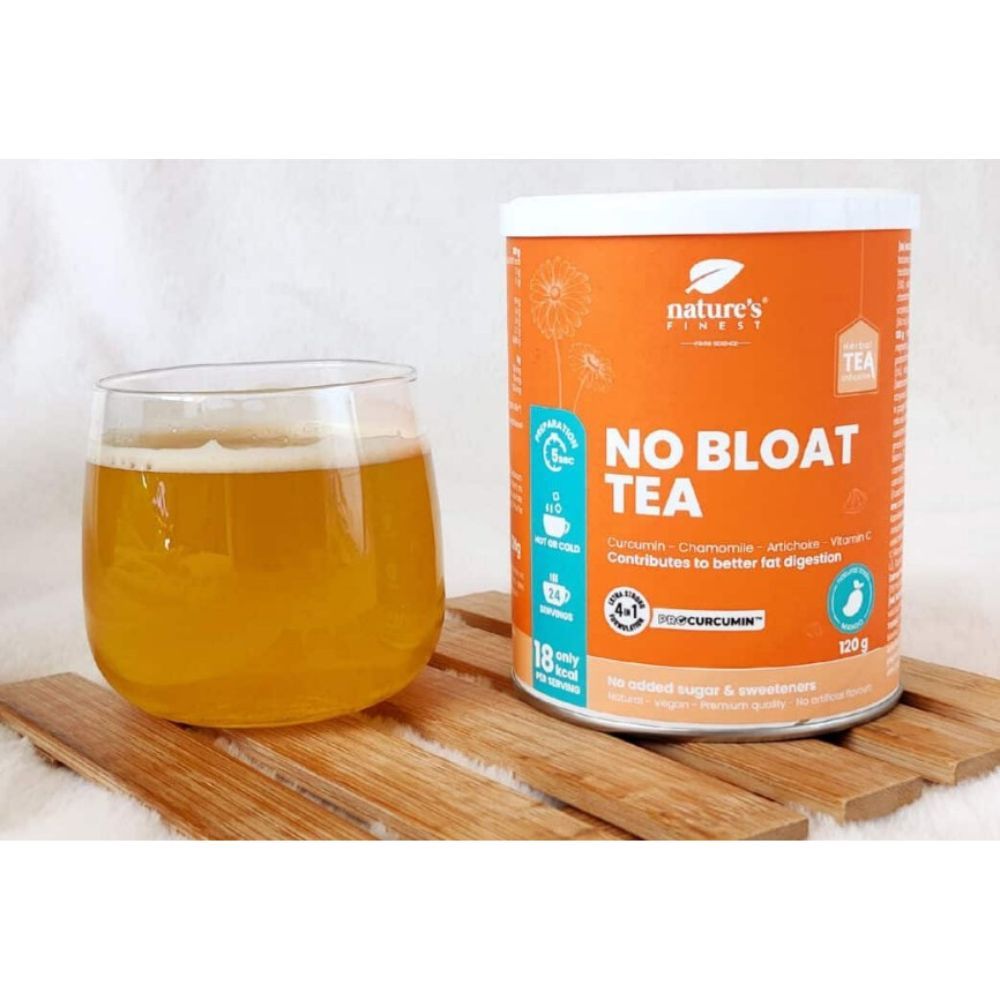 Glas Tee neben Dose "Nature's Finest No Bloat Tea". Dose orange, Etikett mit Zutaten. Tee goldfarben, in Glas. Holzuntergrund.