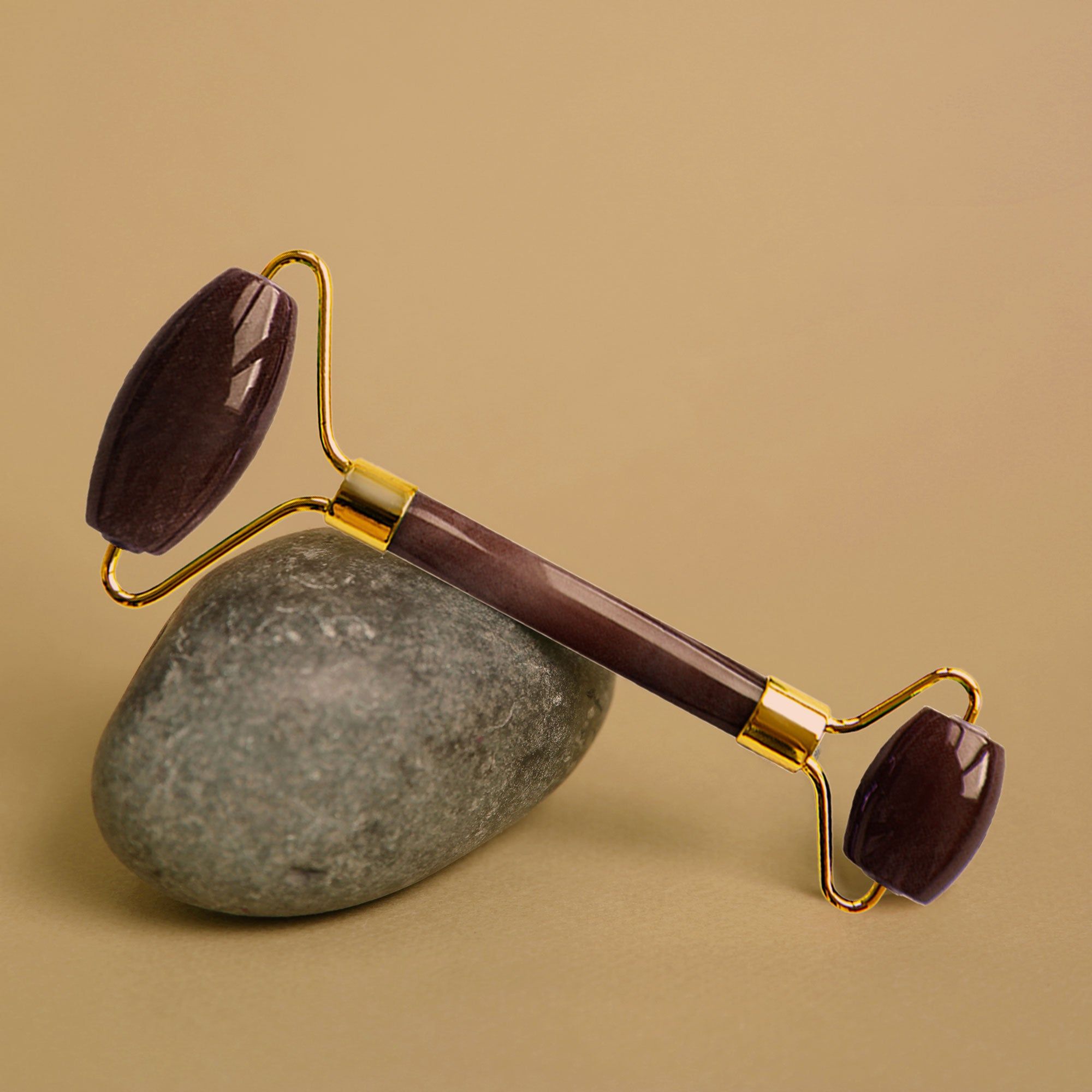 Gesichtsroller auf einem Stein. Der Roller hat einen goldenen Griff und braune Rollen. Der Stein ist grau und der Hintergrund beige.