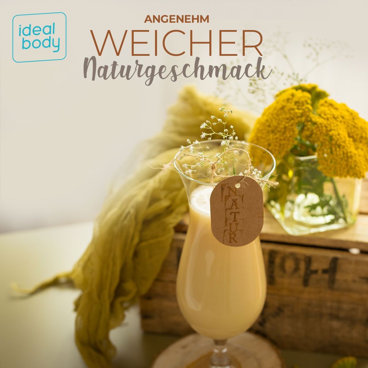 Glas mit hellem Proteinshake, dekoriert mit Blumen. Aufschrift: Angenehm Weicher Naturgeschmack. Auf Holzbrett und Holzkiste.