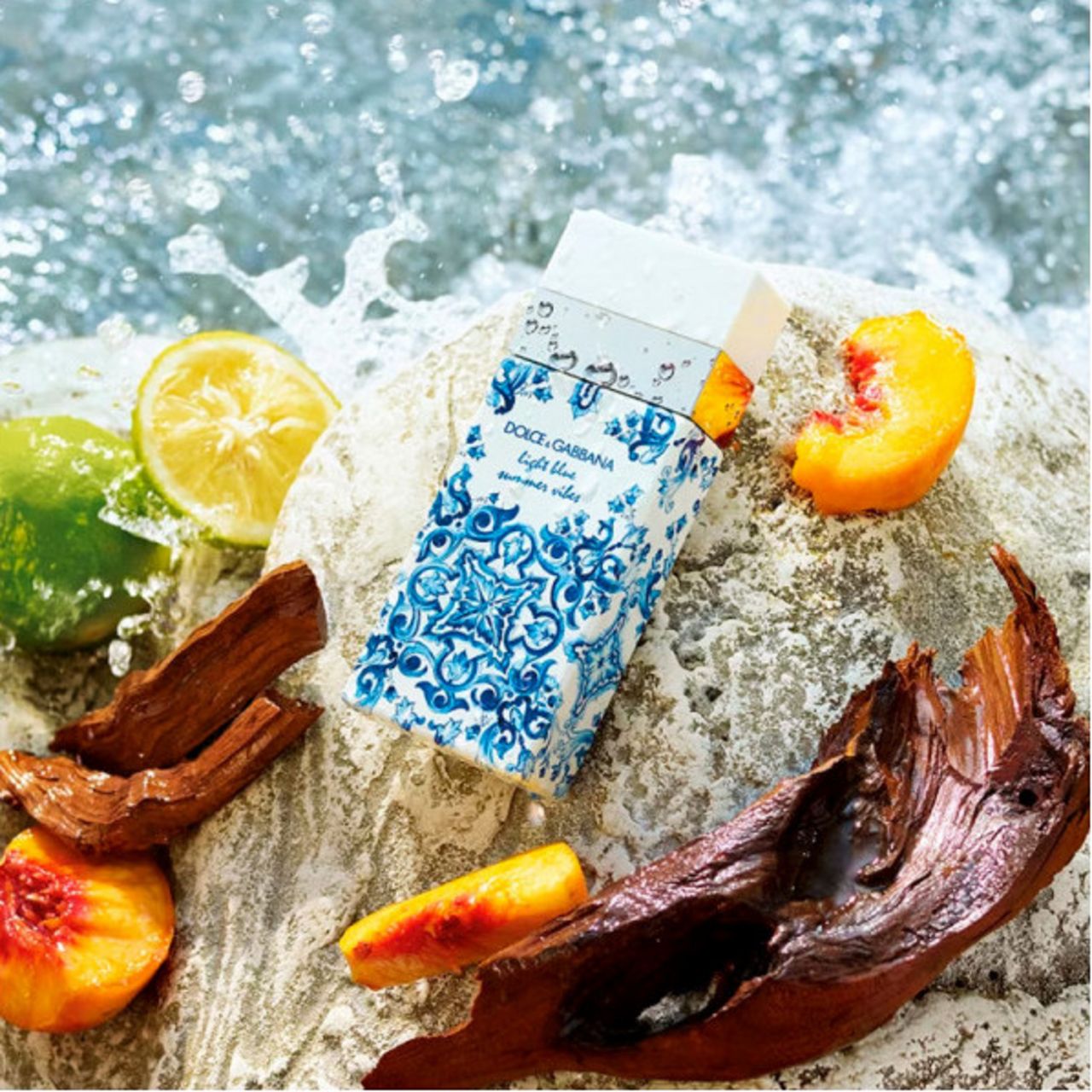 Parfumflakon auf Stein. Früchte und Holzstücke. Aufschrift: Dolce & Gabbana Light Blue Summer Vibes. Spritzwasser.