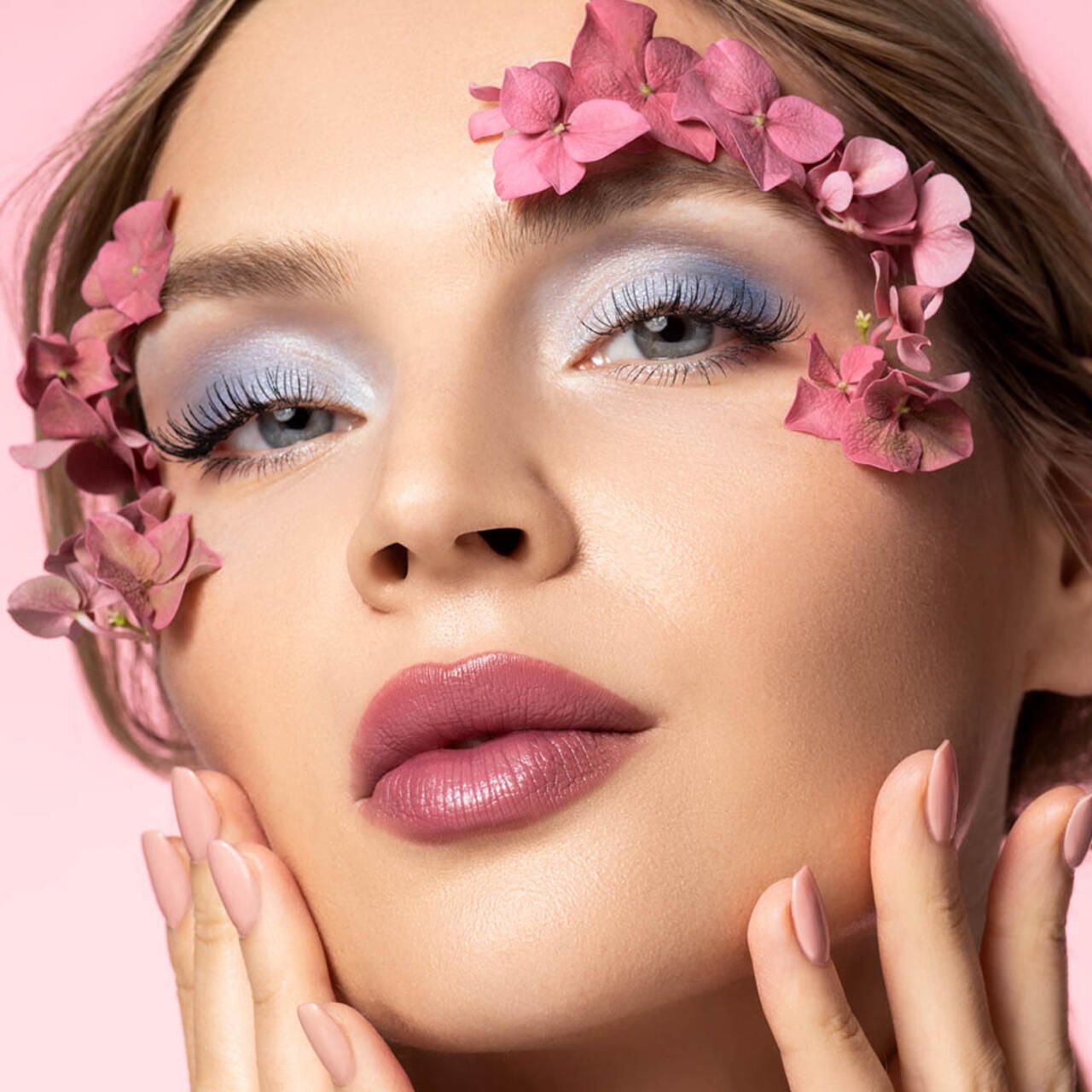 Porträt einer Frau mit rosa Lippen und Nägeln. Sie trägt Lidschatten und Blüten.
