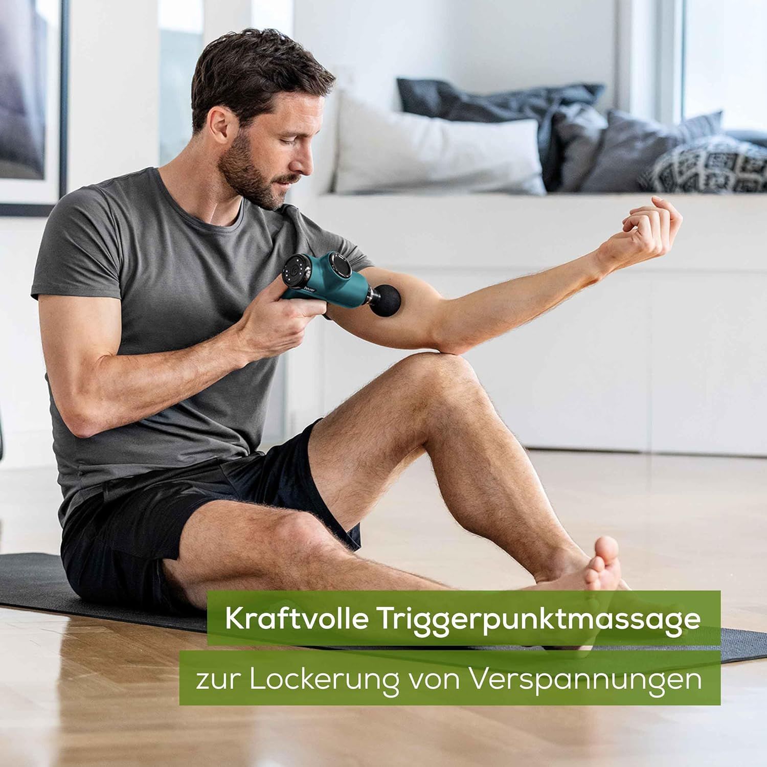 Person sitzt auf Matte und massiert Arm mit Massagepistole. Grün-schwarzes Gerät. Text: Kraftvolle Triggerpunktmassage zur Lockerung von Verspannungen.