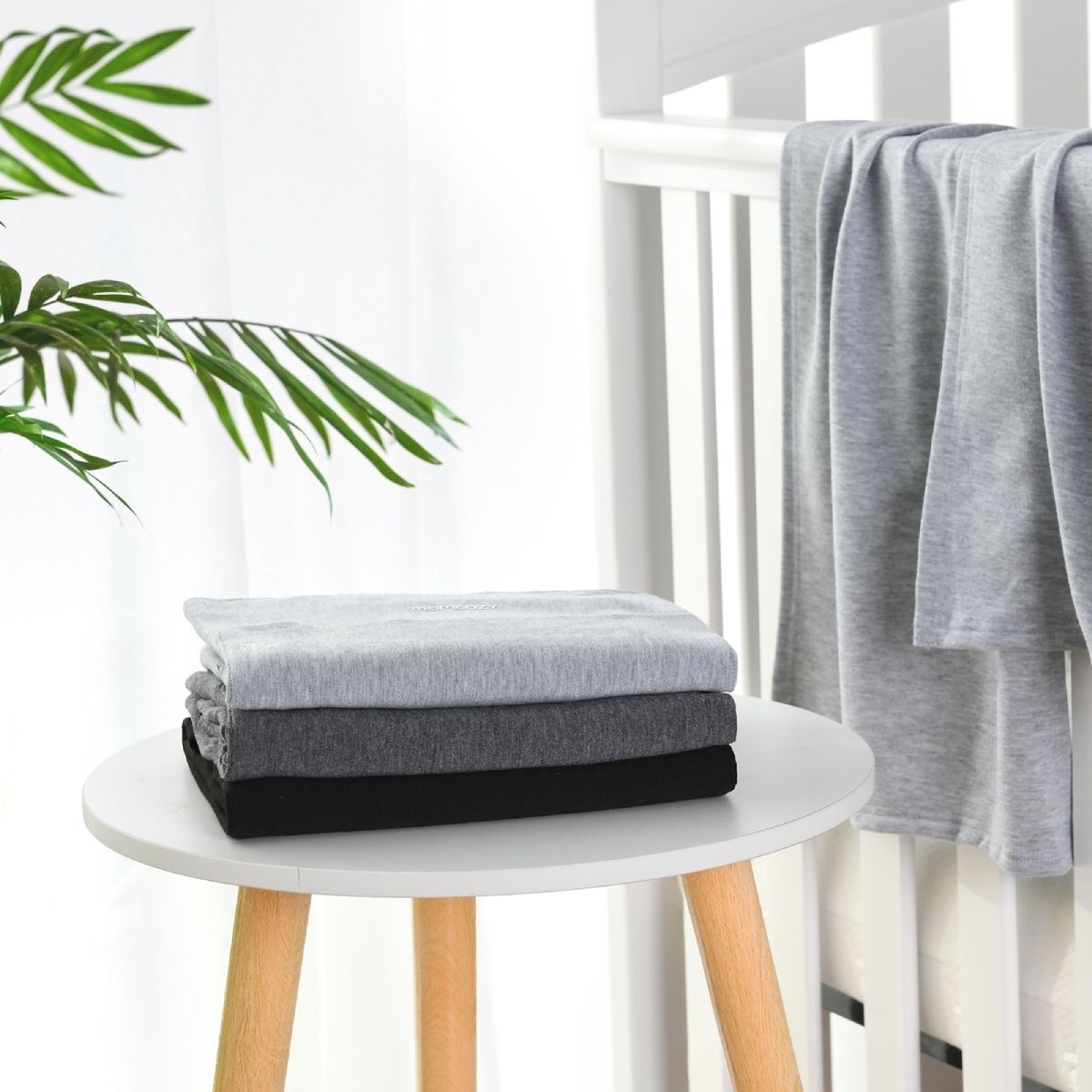 Drei gefaltete Babytragetücher in Grau- und Schwarztönen, gestapelt auf einem kleinen weißen Tisch. Ein weiteres Tuch hängt über der Lehne eines weißen Kinderbettes. Im Hintergrund sind grüne Blätter zu sehen.