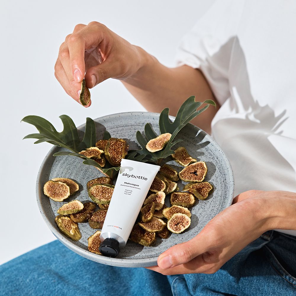 Person hält eine getrocknete Feige über einer Schale mit Feigen und Blättern. Eine Tube liegt in der Schale.