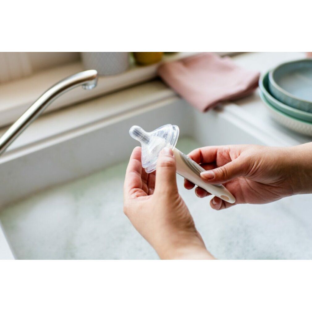 Hände halten eine transparente Brustwarze und einen Flaschenhals über einem Waschbecken. Im Hintergrund ein Wasserhahn und Geschirr.