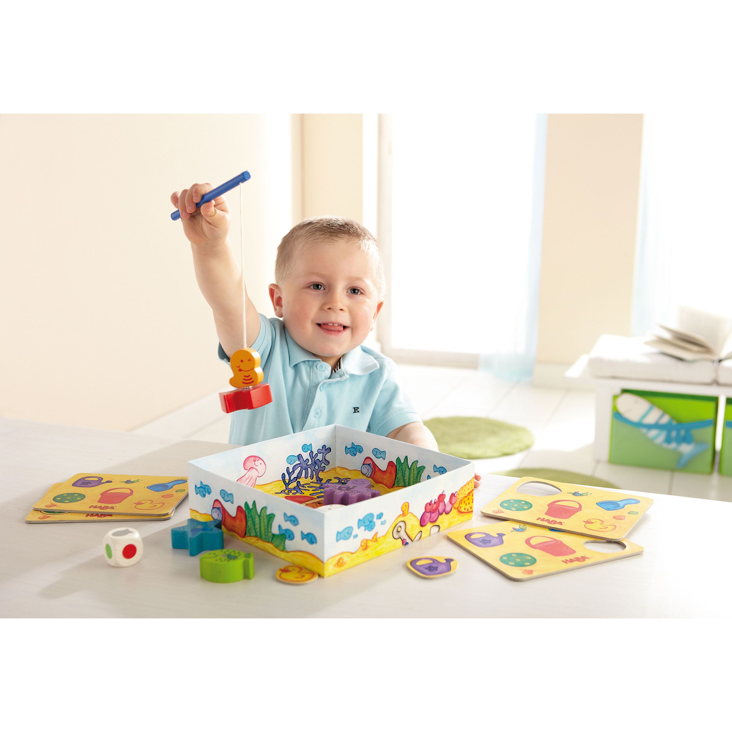 Kind spielt mit "Meine ersten Spiele - Fische angeln". Kind hält Angel über Spielbox mit Fischen. Spielzubehör.