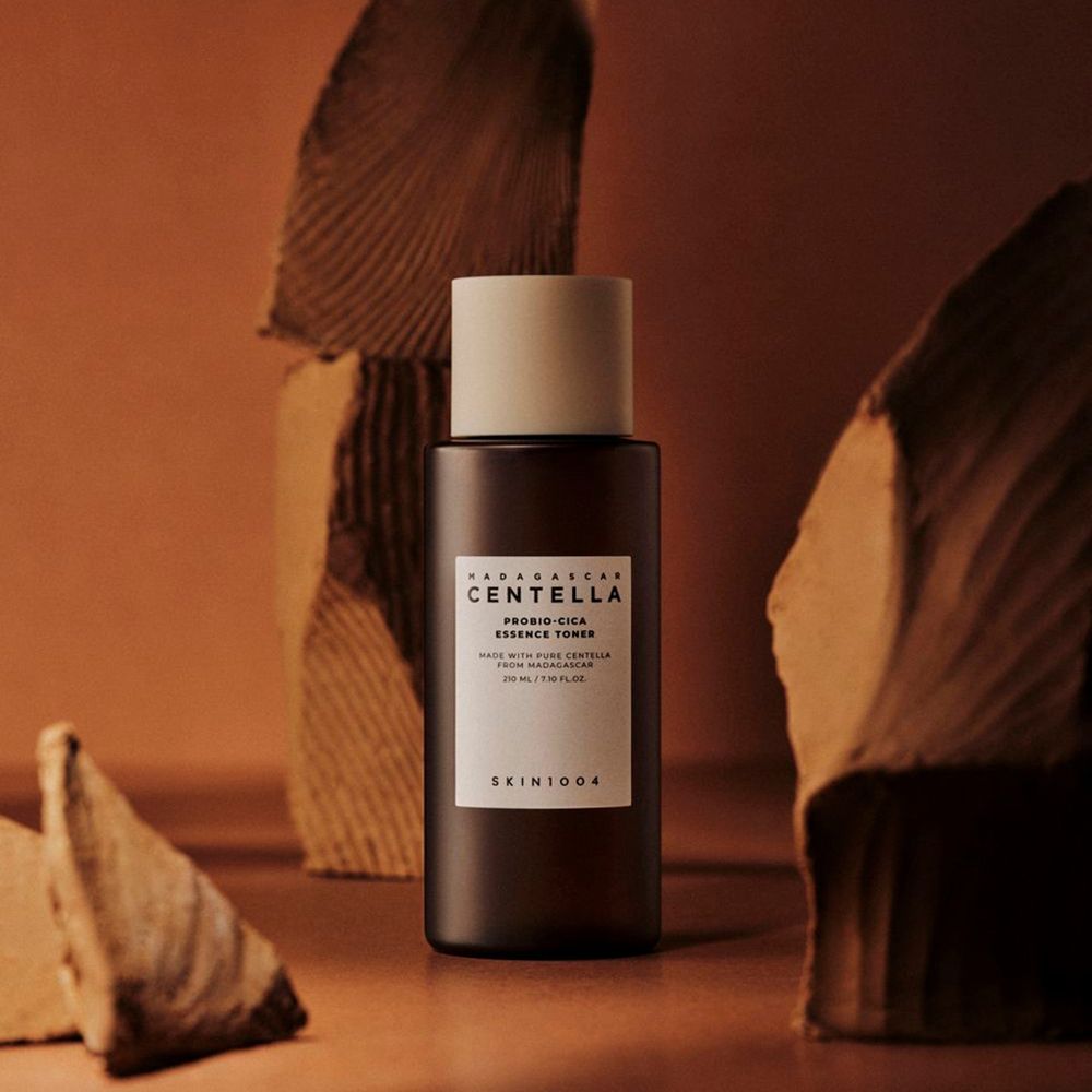 Braune Flasche mit weißem Deckel. Aufkleber mit Text: CENTELLA, PROBIO-CICA ESSENCE TONER, SKIN1004. Vor braunem Hintergrund.