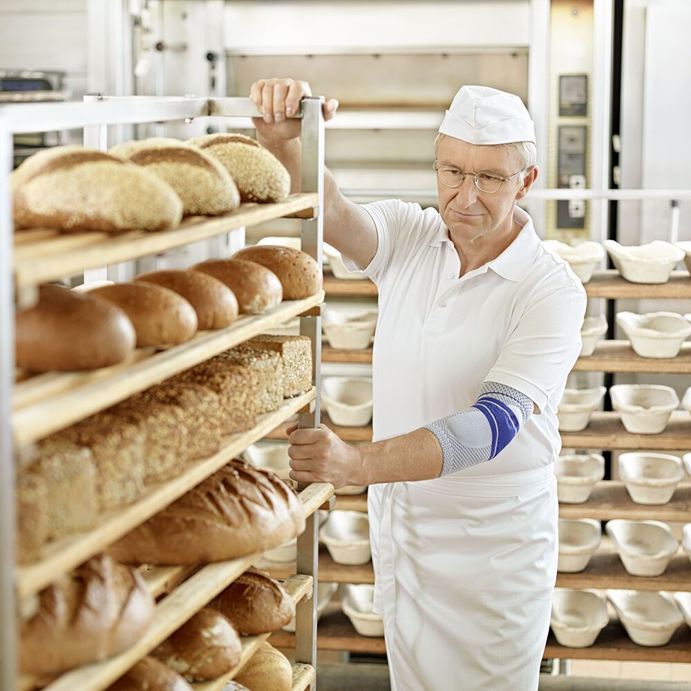 Bäcker mit Ellenbogenbandage vor Regalen mit Brot. Er schiebt einen Wagen.