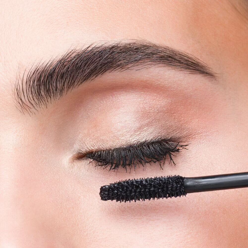 Auge mit Wimpern, die mit Mascara getuscht werden. Mascarabürste in der Nähe.