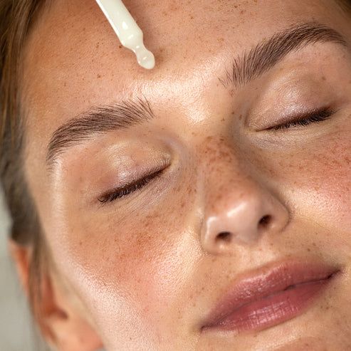 Gesicht einer Frau mit geschlossenen Augen. Eine Pipette tropft Serum auf die Stirn. Haut mit Sommersprossen.