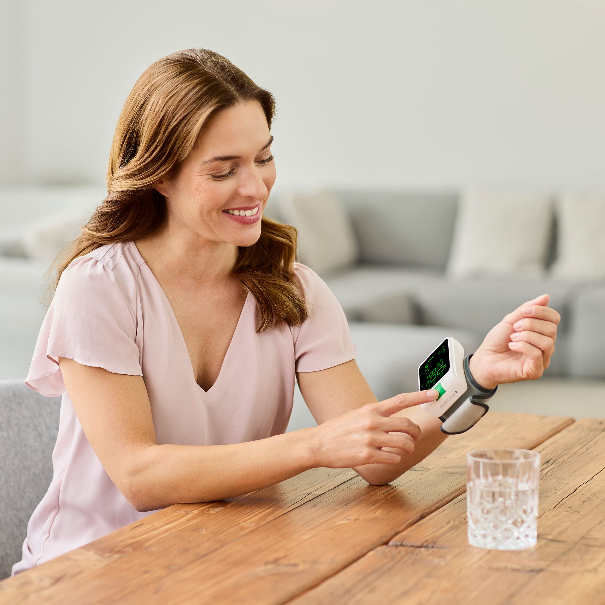 Frau misst Blutdruck am Handgelenk mit einem medisana Gerät. Ein Glas Wasser steht auf dem Tisch.
