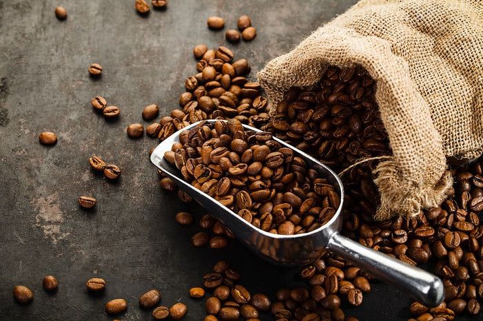 Kaffeebohnen, teilweise aus einem Jutesack, mit silberner Schaufel. Bohnen auf dunklem Untergrund.