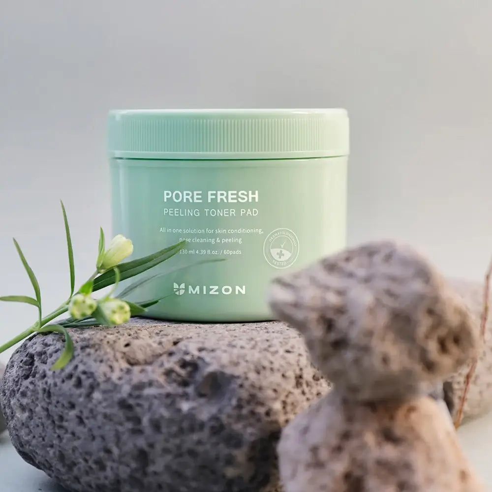 Grüne Dose mit Toner-Pads auf Stein. Aufschrift: Pore Fresh Peeling Toner Pad. Marke: MIZON. Mit Blüte.