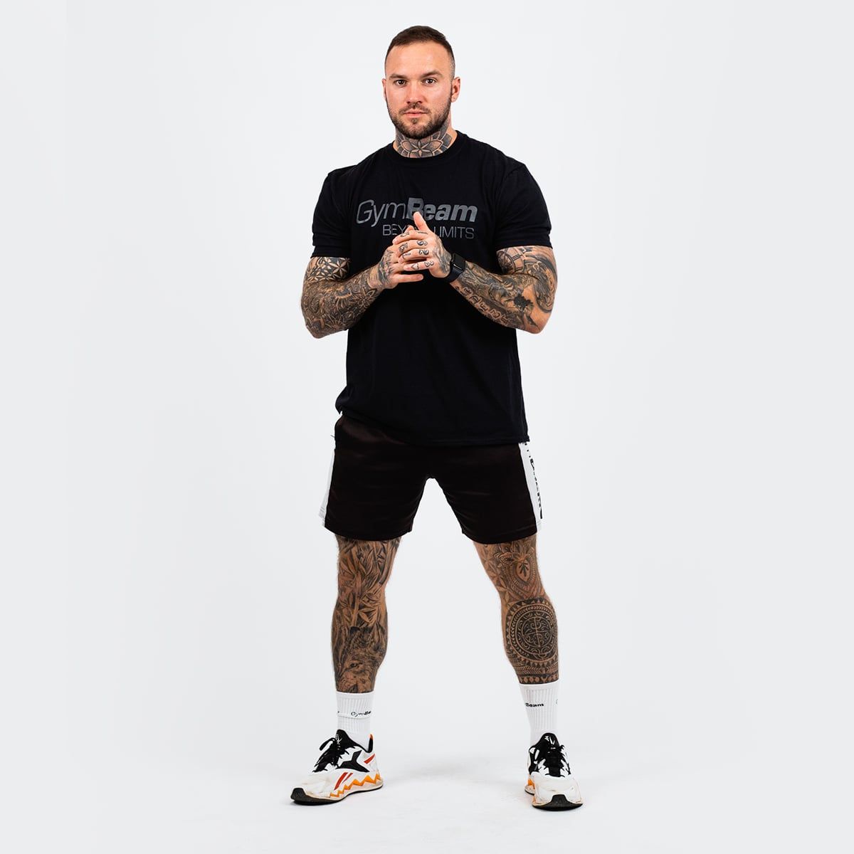 Ein Mann in voller Länge trägt ein schwarzes GymBeam T-Shirt und schwarze Shorts. Er steht vor einem weißen Hintergrund.