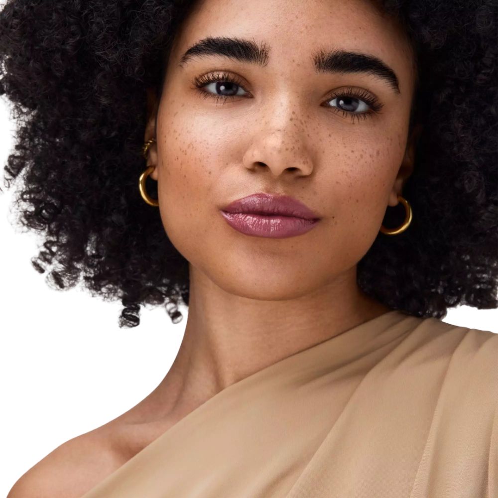 Porträt einer Frau mit Locken und rosafarbenen Lippen. Sie trägt goldene Ohrringe und ein beiges Oberteil.