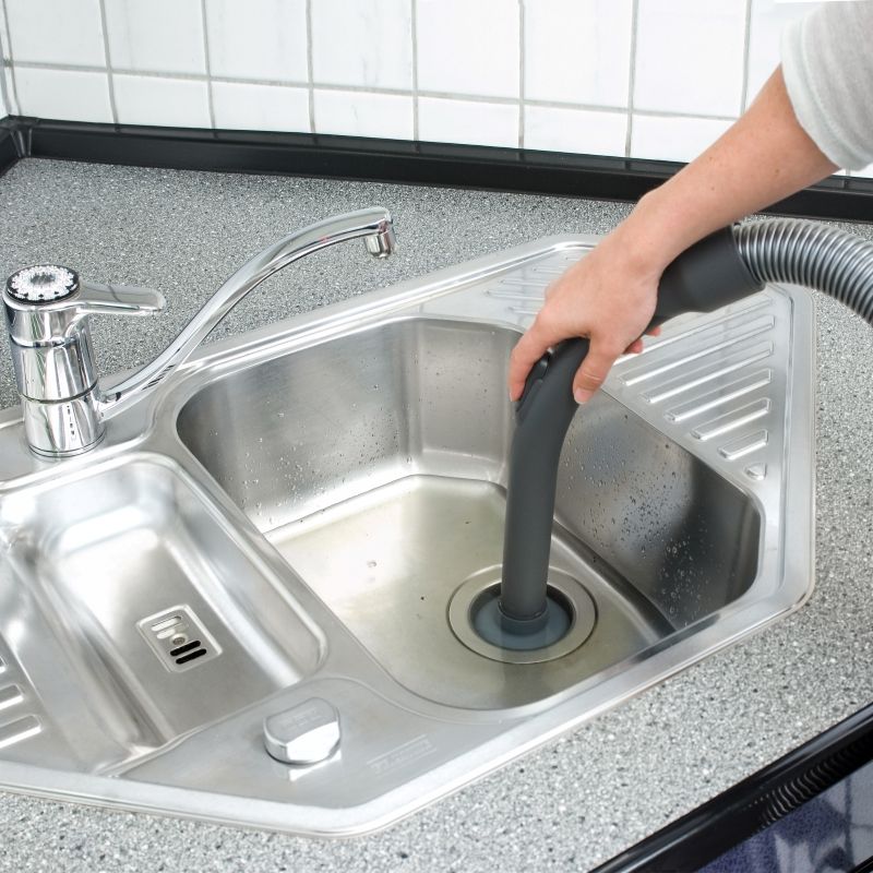 Hand mit Schlauch in Spülbecken. Edelstahlbecken, grauer Schlauch, Wasserhahn.