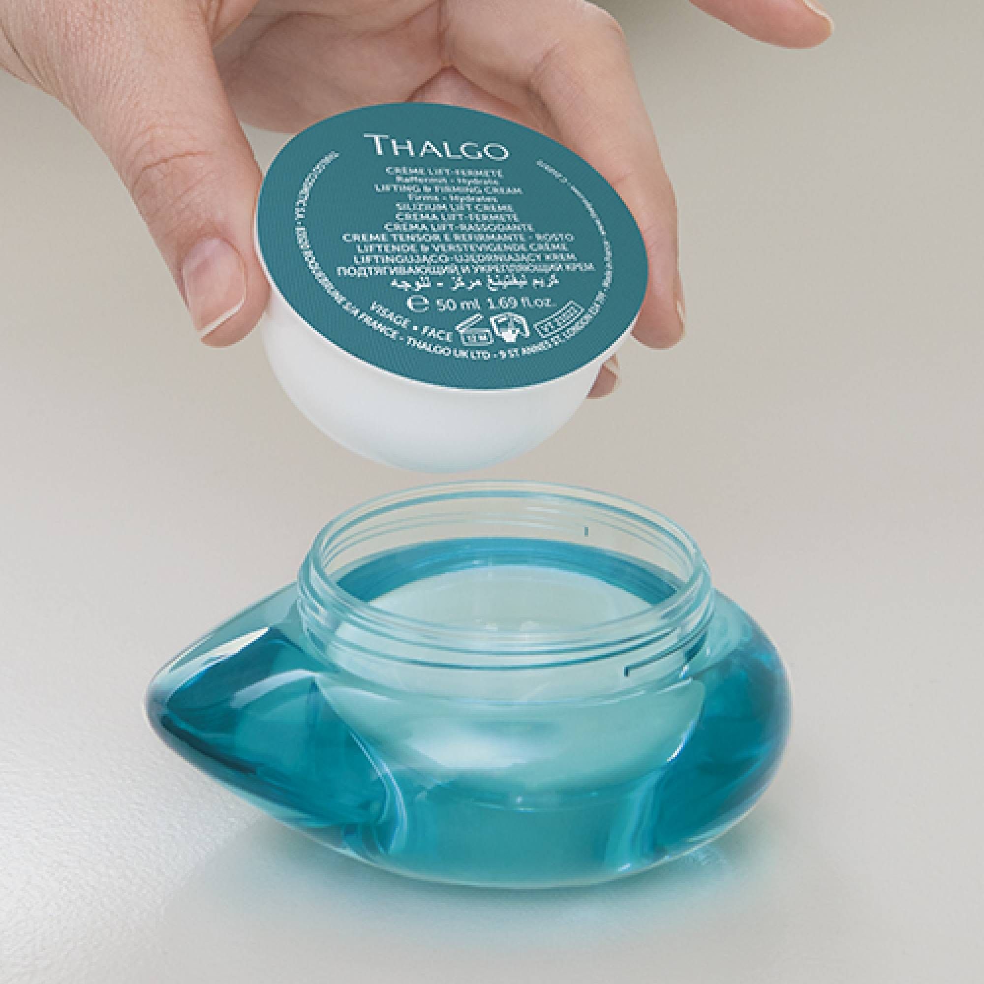 Hand hält eine geöffnete Creme-Verpackung über einem leeren, blauen Glasbehälter. Aufschrift: THALGO.