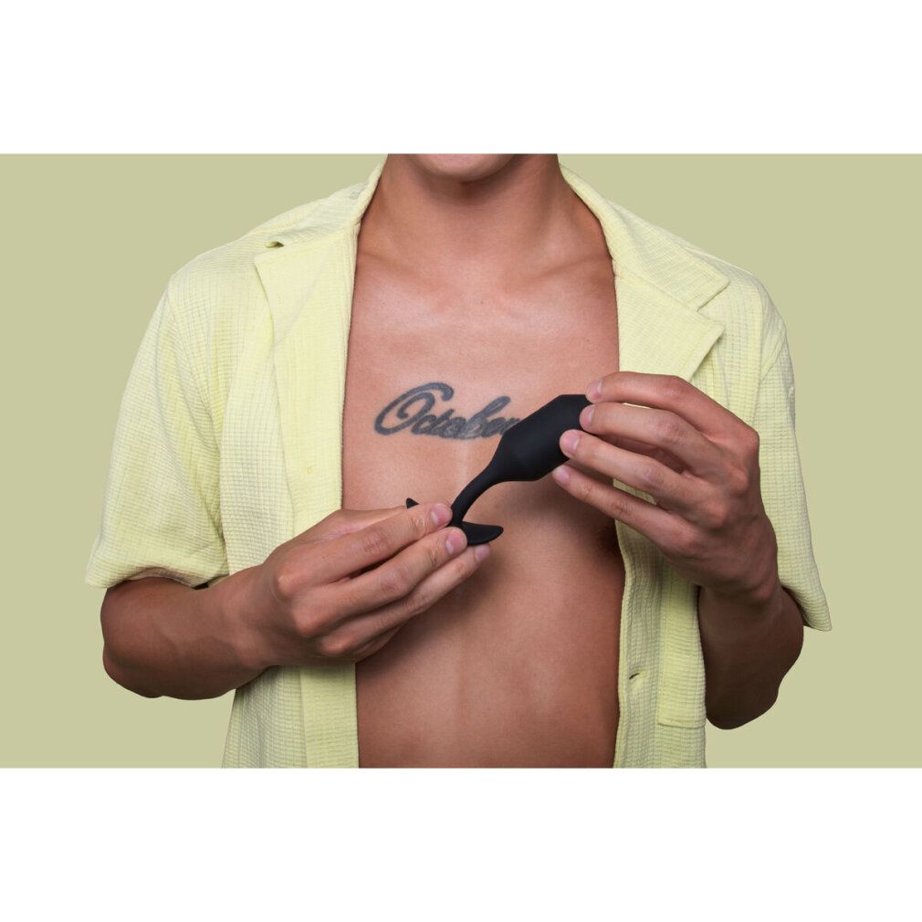 Person hält schwarzen Plug. Brust mit Tattoo 'October'. Gelbes Hemd.