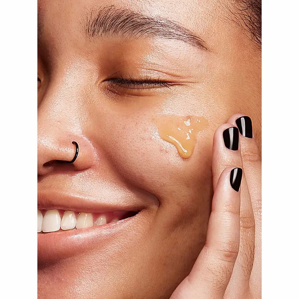 Person mit Produkt auf der Haut. Gelbliche Creme auf der Wange. Schwarze Fingernägel.