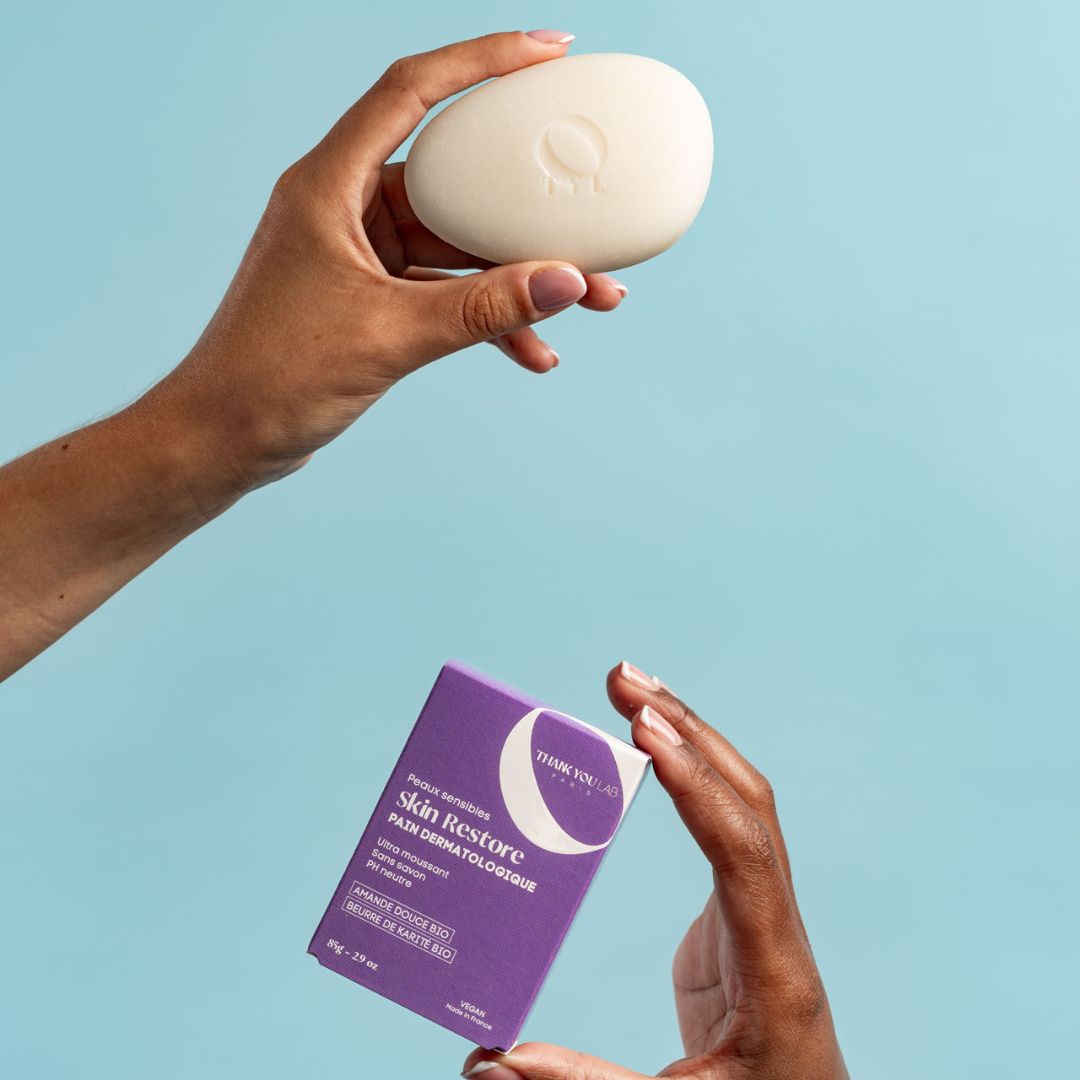 Hände halten ein ovales Seifenstück und eine lila Schachtel. Auf der Schachtel steht "Skin Restore".