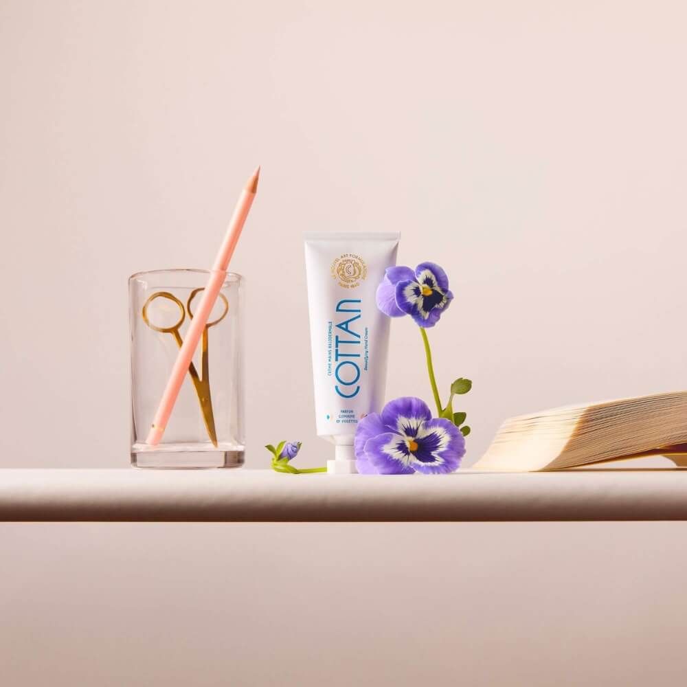 Weiße Tube mit blauem Schriftzug COTTAN, daneben Stiefmütterchen, Bleistift, Schere und Buch.