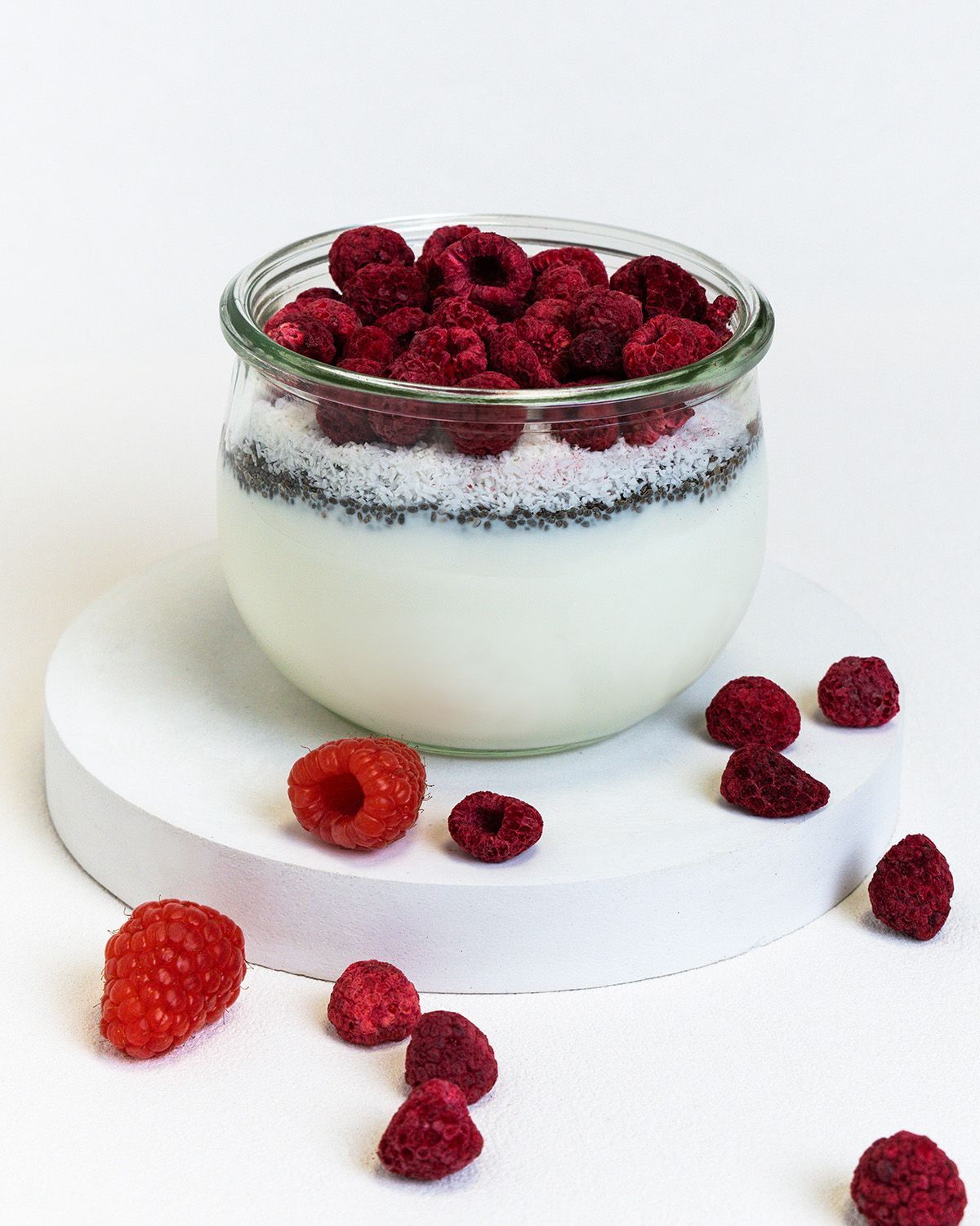 Glas mit Joghurt, Chia-Samen und Himbeeren. Einige Himbeeren liegen verstreut auf einer weißen Oberfläche.