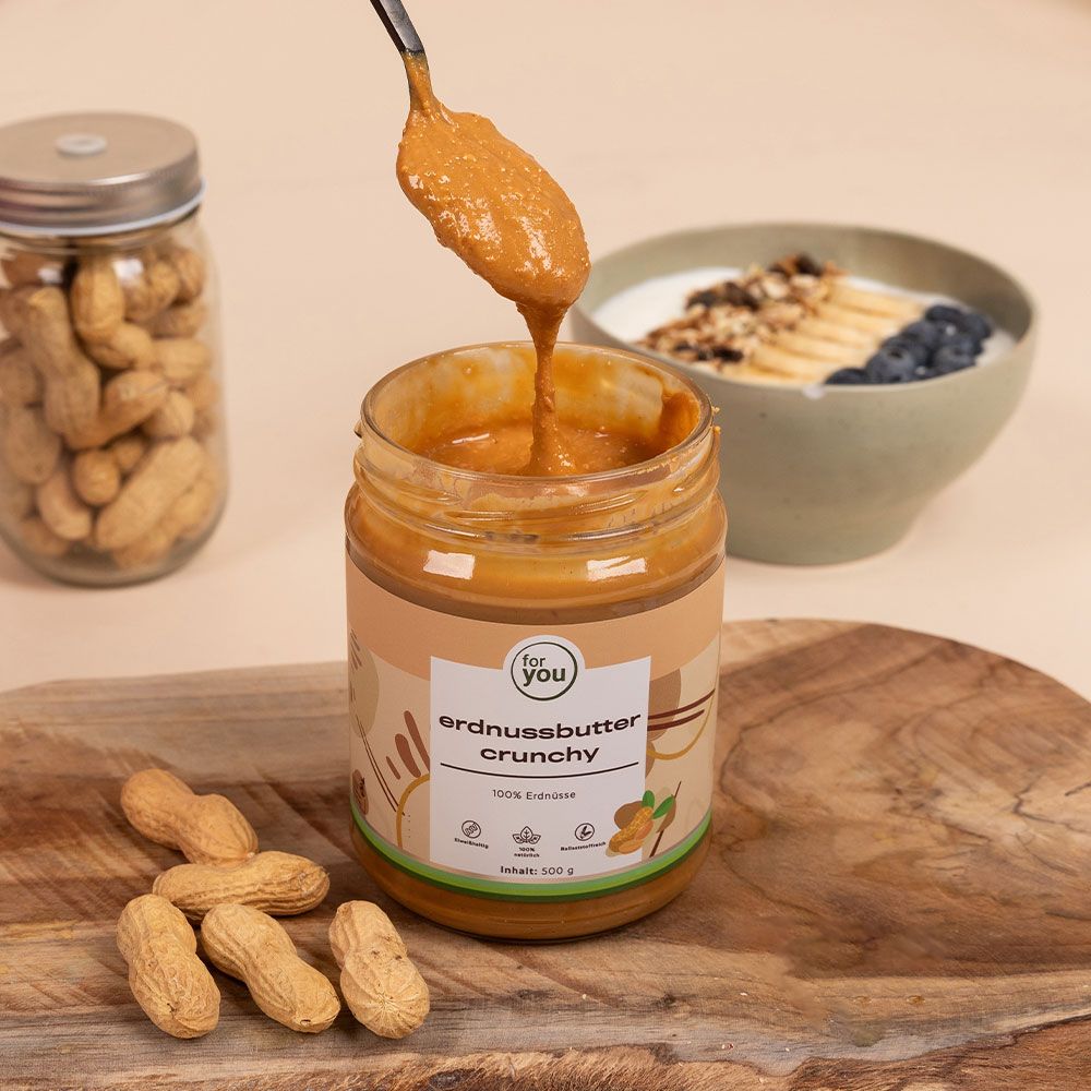 Glas Erdnussbutter Crunchy, Löffel mit Butter, Schüssel mit Joghurt, Banane und Blaubeeren, Erdnüsse. Aufschrift: for you, erdnussbutter crunchy, 100% Erdnüsse. Inhalt: 500 g.