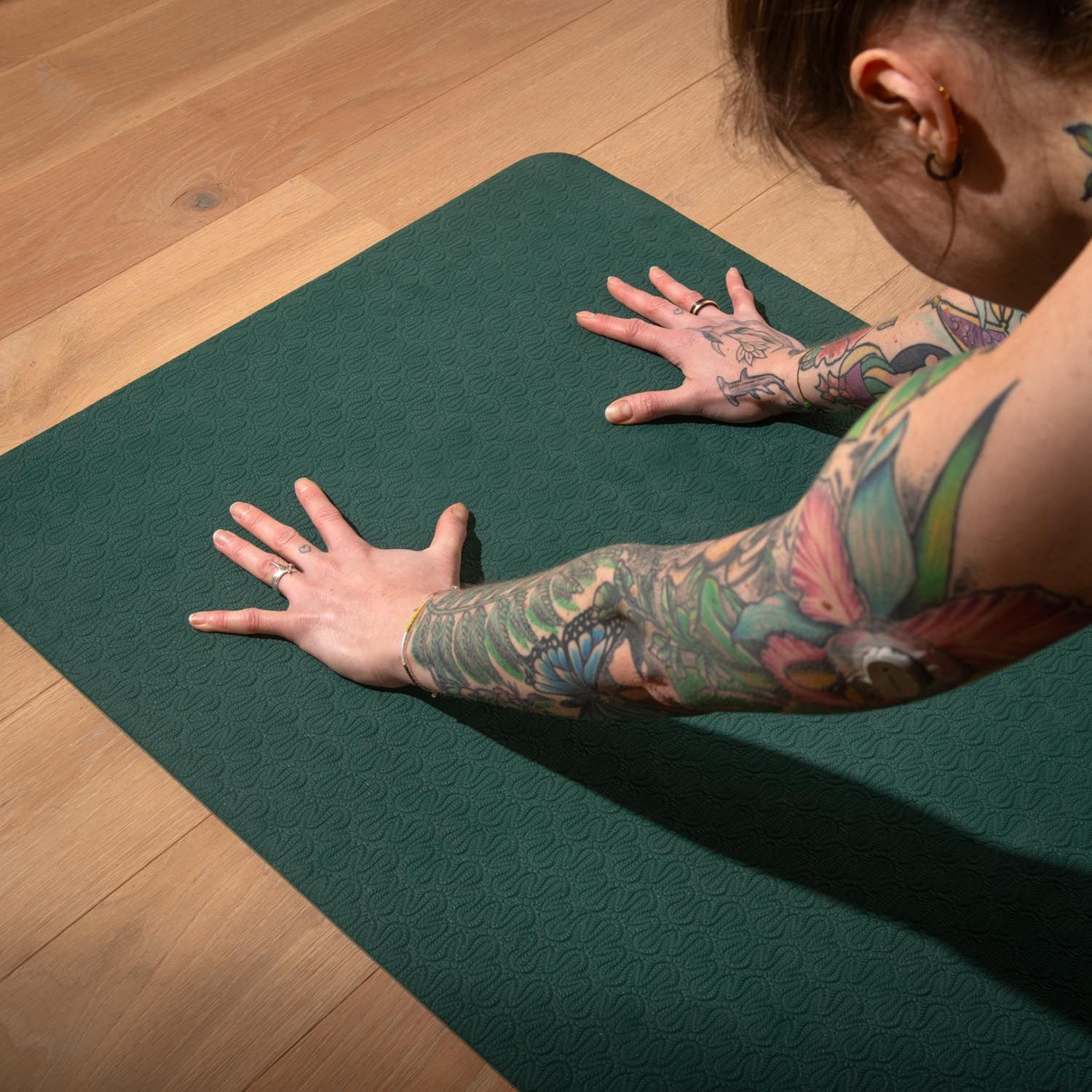 Person liegt auf einer dunkelgrünen Yogamatte. Arme und Hände sind ausgestreckt. Holzfußboden im Hintergrund.