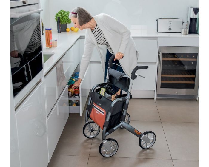 Rollator mit Einkaufstasche. Grauer Rahmen, schwarze Tasche mit roter Front. Person in Küche. "Trust" Schriftzug.