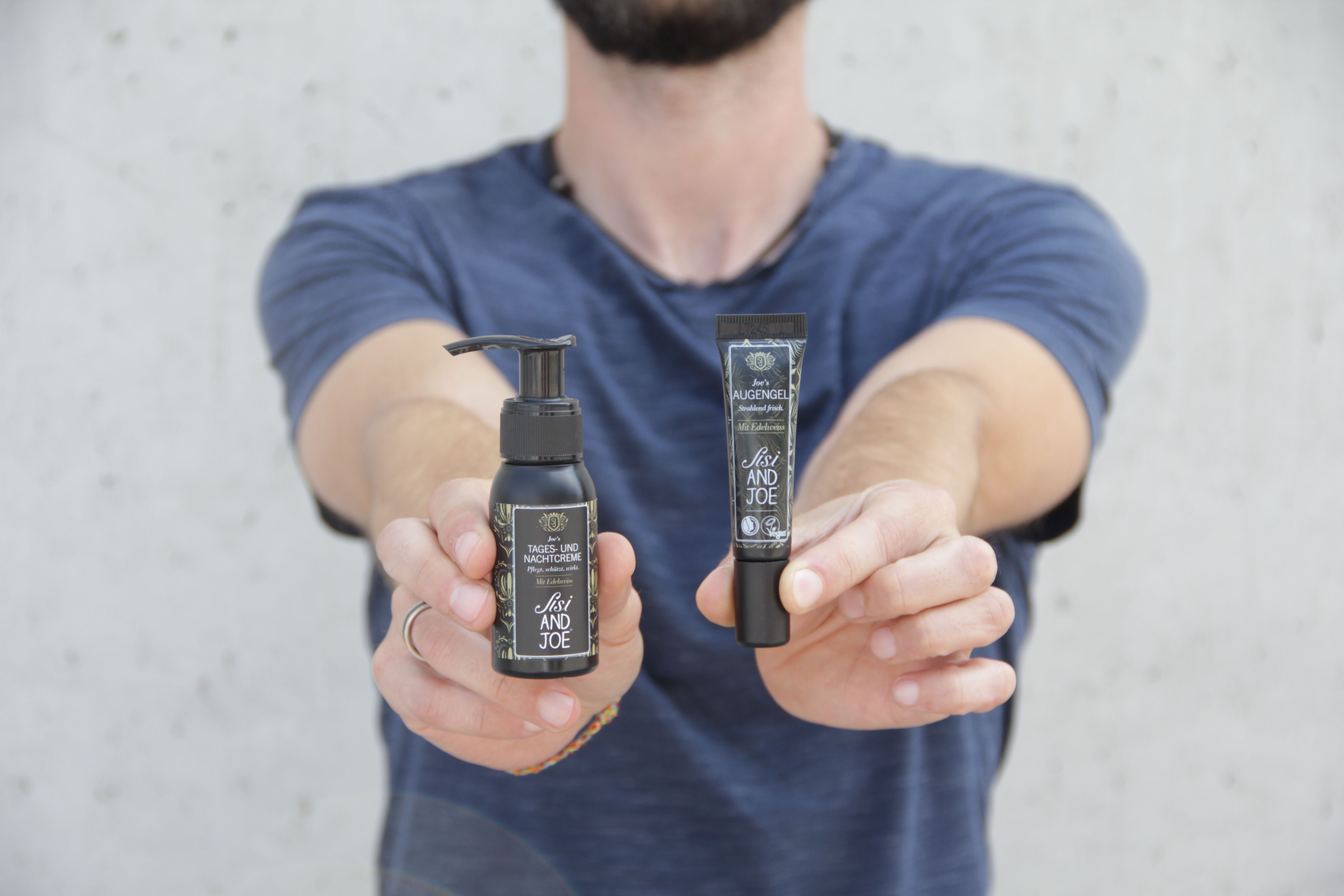 Mann hält zwei schwarze Flaschen. Links: Flasche mit Pumpe. Rechts: Tube. Beide mit Etiketten.