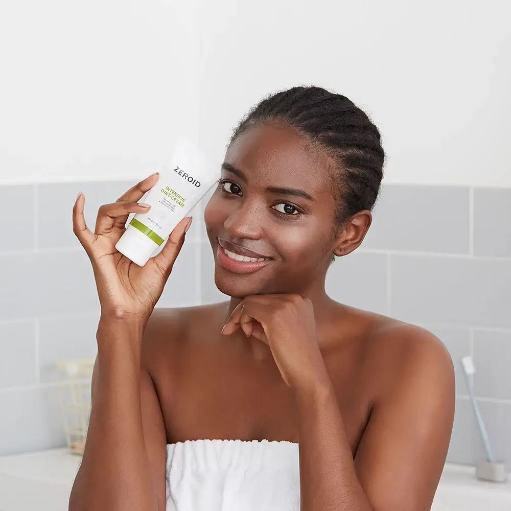 Frau hält Tube ZEROID INTENSIVE OINT-CREAM. Sie lächelt. Hintergrund: Badezimmer mit Fliesen.