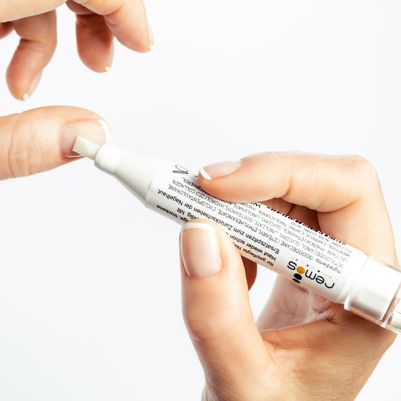 Weißer Stift mit Spitze, wird auf einen Fingernagel aufgetragen. Hände halten den Stift. Aufschrift: remos.