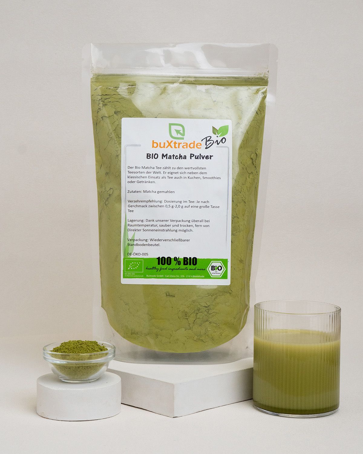 Transparente Verpackung mit Matcha-Pulver, Glas mit Getränk und Schälchen mit Pulver. Aufgedrucktes Logo: buXtrade Bio.