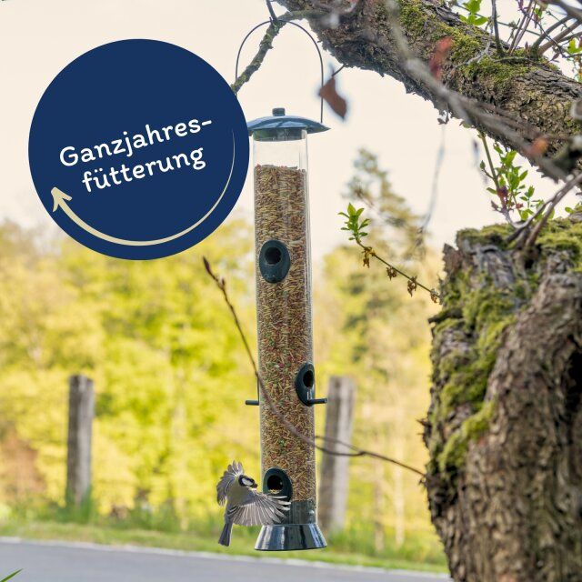 Vogelfutterspender mit Futter. Beschriftung: Ganzjahresfütterung. Vogel fliegt zum Spender.
