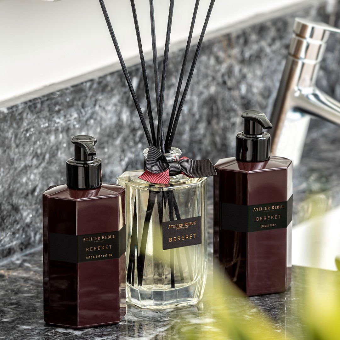 Zwei bordeauxfarbene Lotionflaschen mit schwarzem Pumpkopf. Aufschrift: Atelier Rebul Bereket Hand & Body Lotion. Daneben Duftdiffusor.