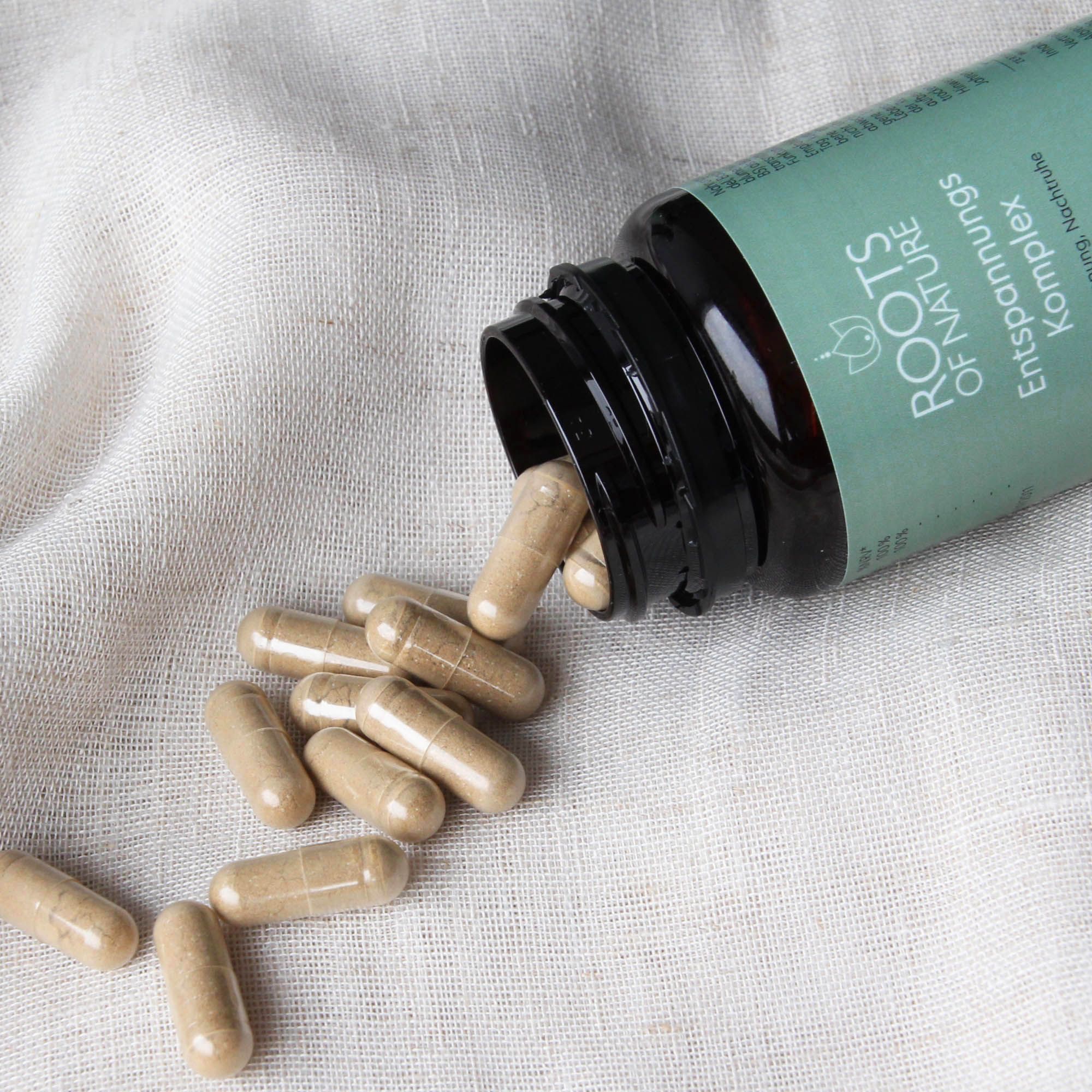 Braune Kapseln liegen neben einer geöffneten Flasche. Produkt: ROOTS OF NATURE Entspannungs Komplex. Auf hellem Stoff.