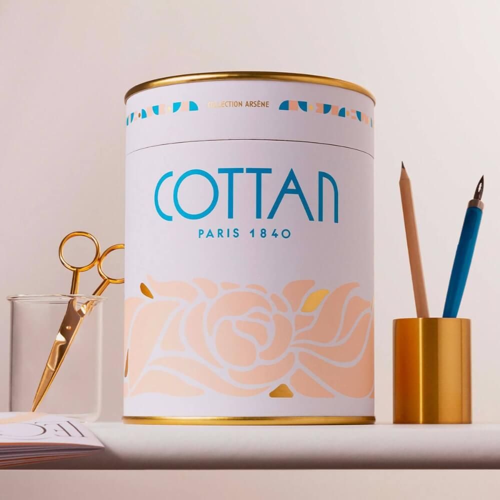 Zylindrische Dose mit goldfarbenem Deckel und Boden. Aufschrift COTTAN PARIS 1840. Schere, Stifte und Becher daneben.