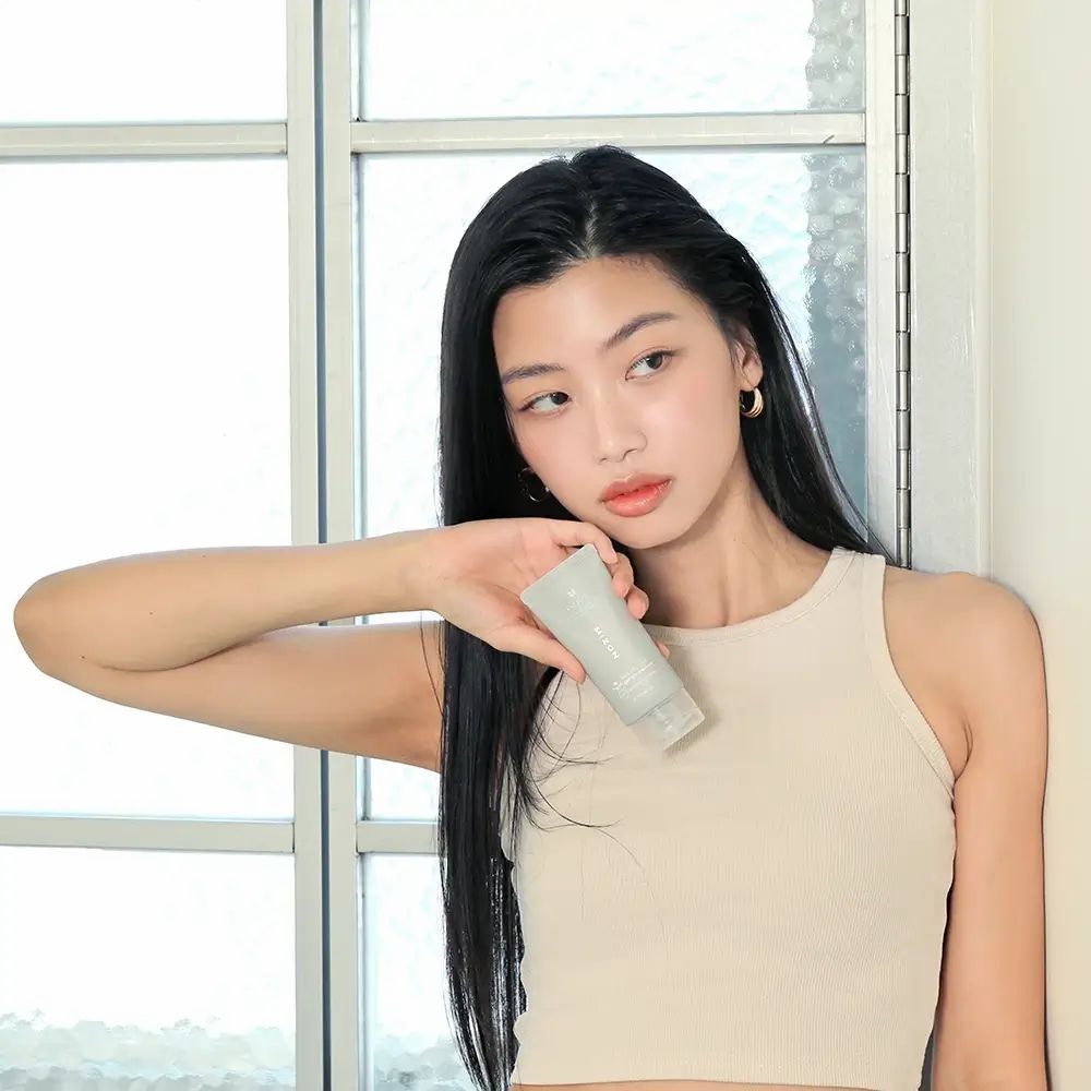 Junge Frau hält eine graue Tube. Sie trägt ein beiges Top. Hintergrund: Fenster und weiße Wand.