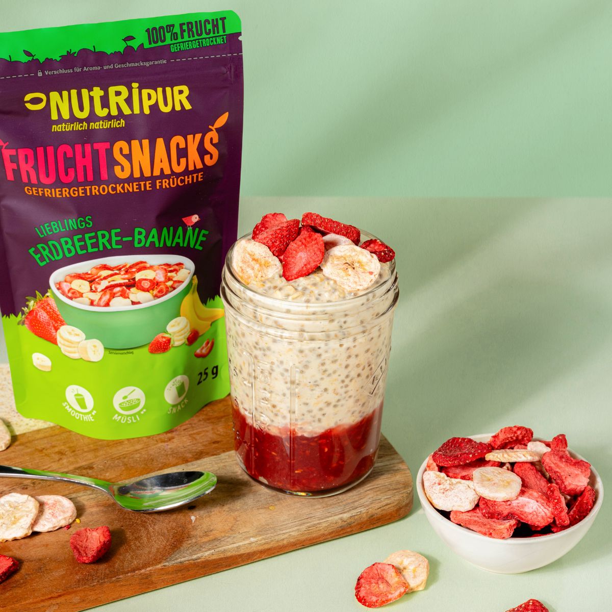 Produktverpackung und ein Glas mit Schichten aus Chia-Pudding und gefriergetrockneten Erdbeeren und Bananen. Schale mit Früchten.
