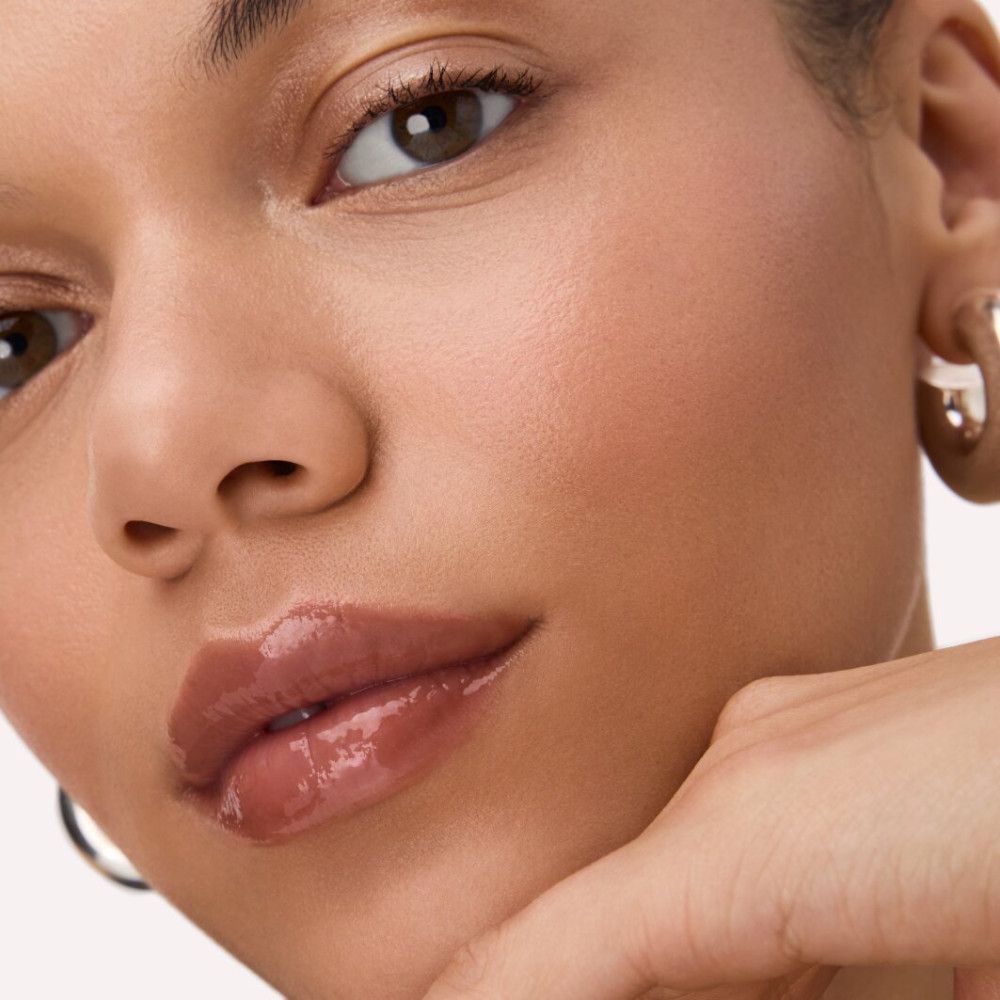 Nahaufnahme eines Gesichts mit glänzendem Lipgloss auf den Lippen. Die Person stützt das Kinn mit der Hand.