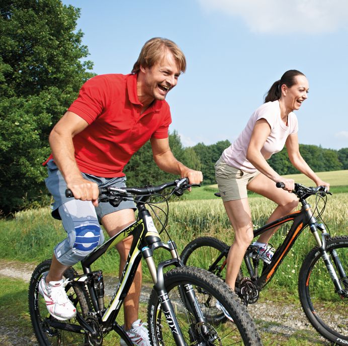 Zwei Personen fahren Fahrrad. Eine Person trägt eine BAUERFEIND GenuTrain S Pro Kniebandage am linken Knie.