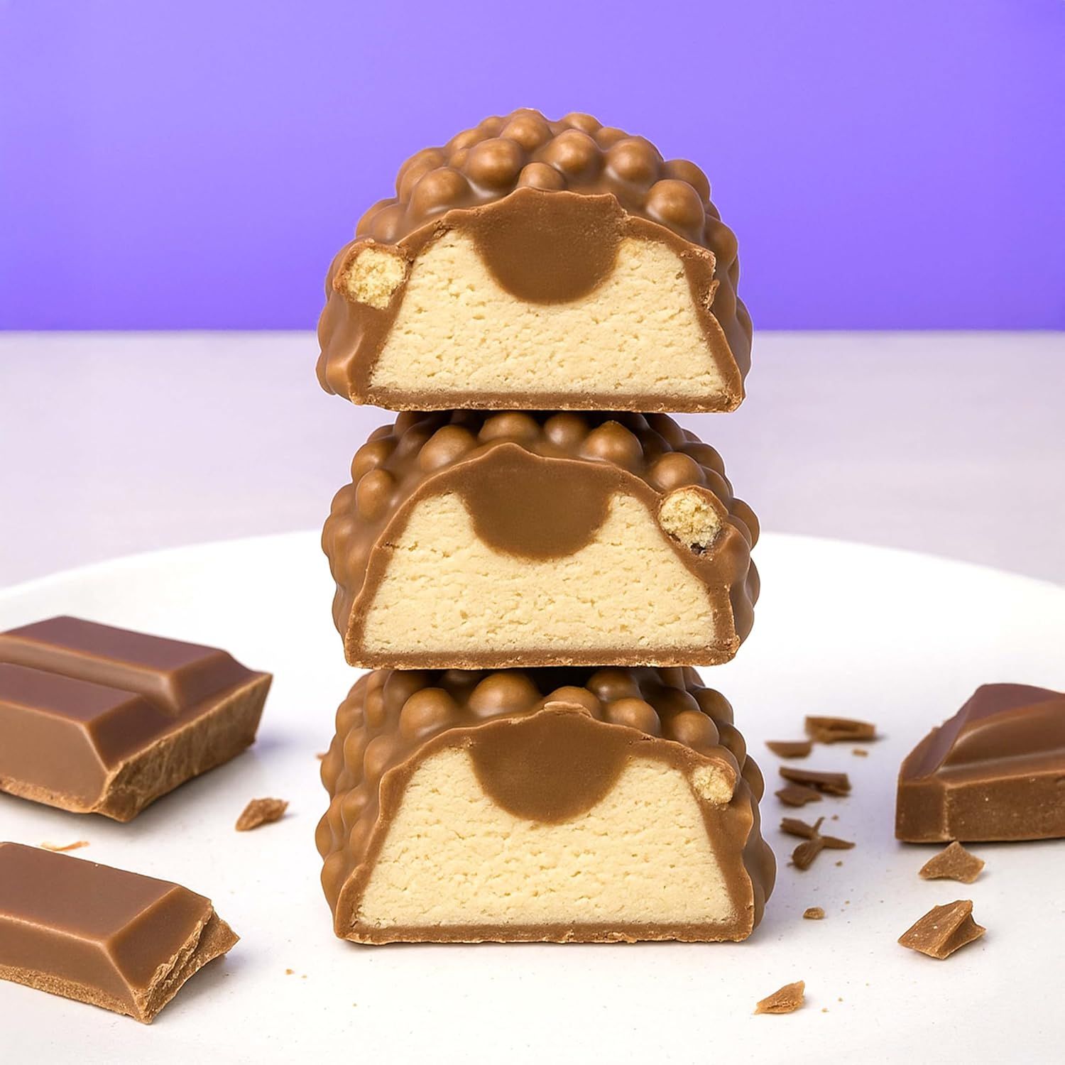 Drei halbierte Proteinriegel, gestapelt. Die Riegel haben eine helle Füllung und eine Schicht aus knusprigen Kugeln. Schokoladenstücke daneben.