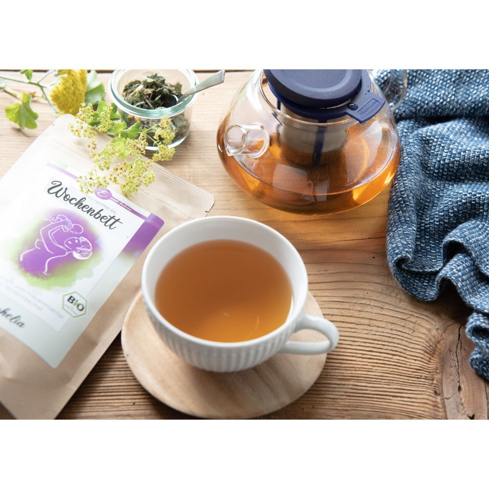 Tasse Tee auf Holzuntergrund. Teebeutel mit Aufschrift Wochenbett. Teekanne. Holzuntersetzer. Blaues Tuch.